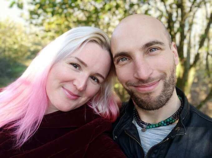 A smiling couple with light hair and a beard, posing closely in a natural outdoor setting, showcasing happiness and connection.