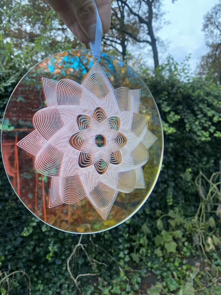 A colorful decorative mandala disc with intricate patterns, suspended by a ribbon against a natural backdrop, showcasing vibrant colors and designs.